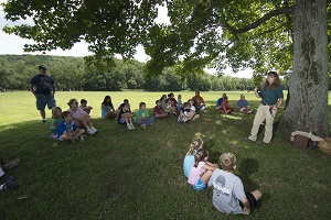 kids sit under the shade of a tree listening to the park naturalist who is holding a snake