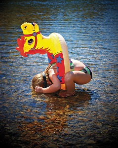 little girl bent over looking into water with goggles on and a horse shaped floaty around her waste