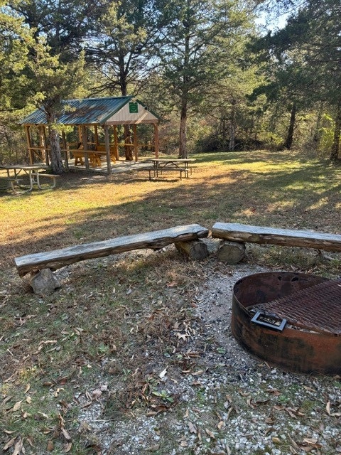 Metal fire pit and rugged wooden benches with wooden picnic tables and open shelter in the background