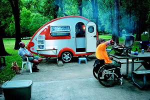 Man in wheel chair cooking over a camp stove with camper and another man in background