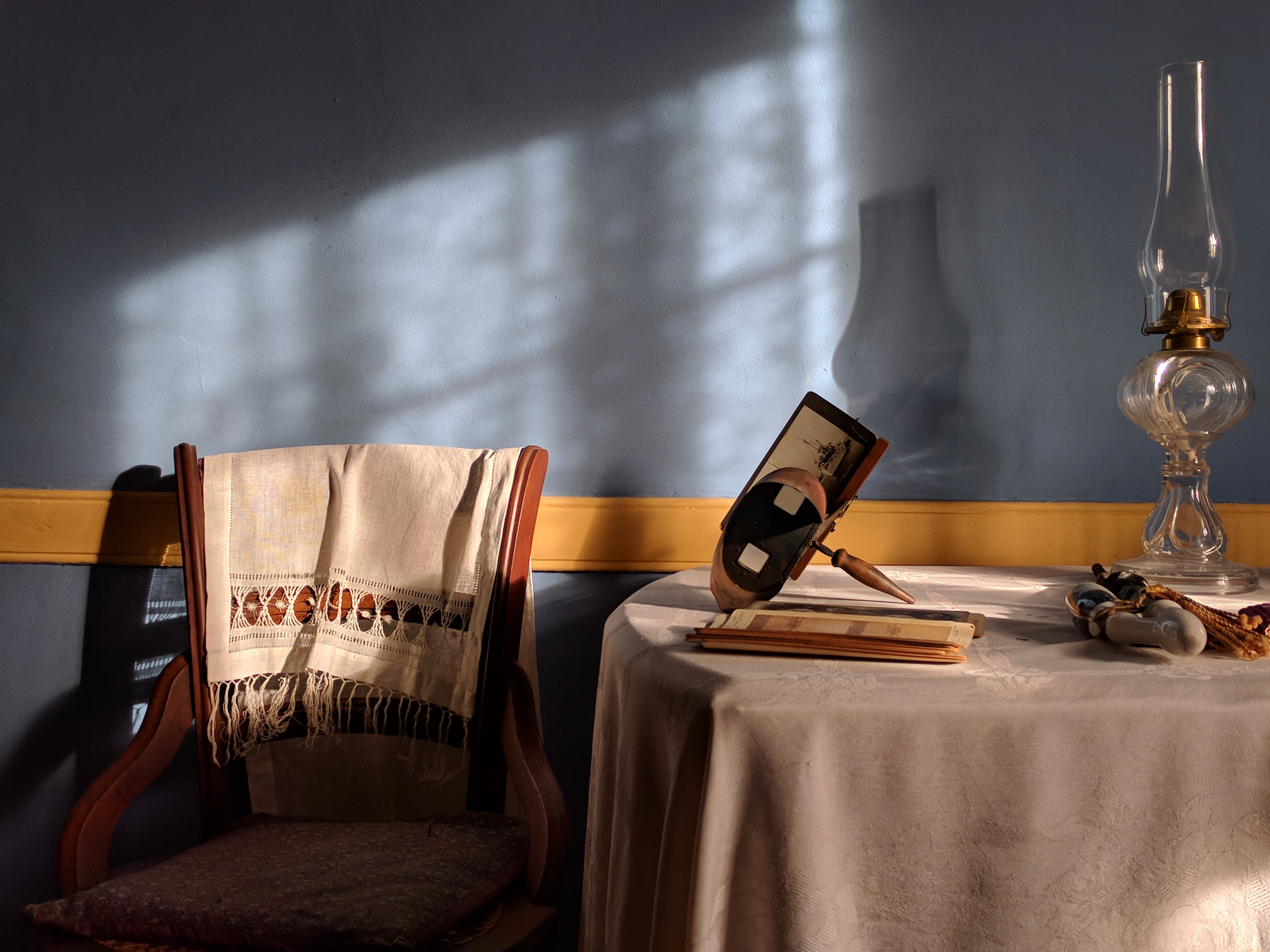 A stereoscope, pipe and oil lamp sit on a table next to a chair with a linen garment strewn over it in a sunlit room