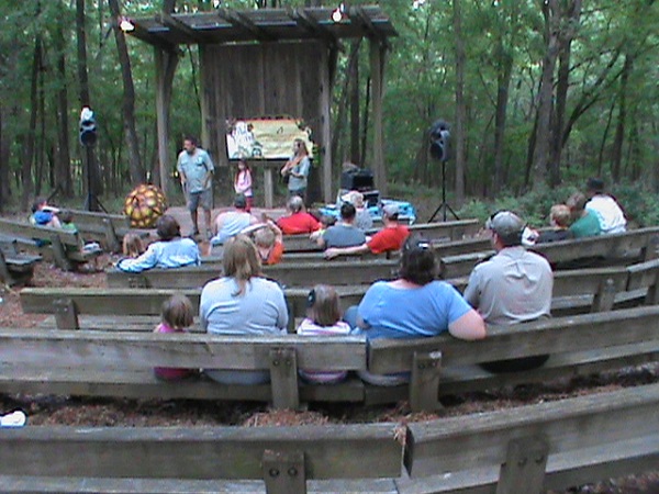 people sit on the wooden benches at the amphitheater listening to a program