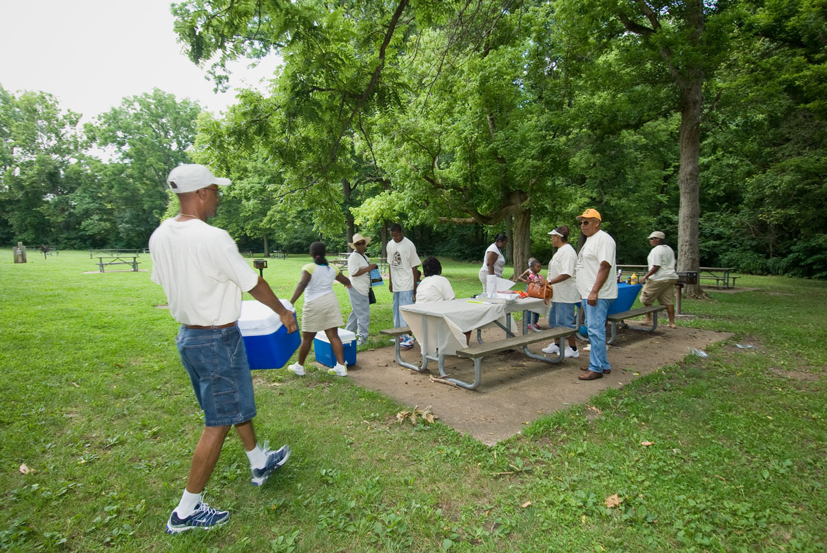 A man carrying a cooler to a picnic table with a family at it