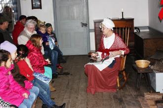 woman in period attire embroidering in front of a group of people