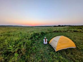 A woman sitting on the ground next to a white and yellow tent looks out across the green grass of the prairie with the sun low on the horizon