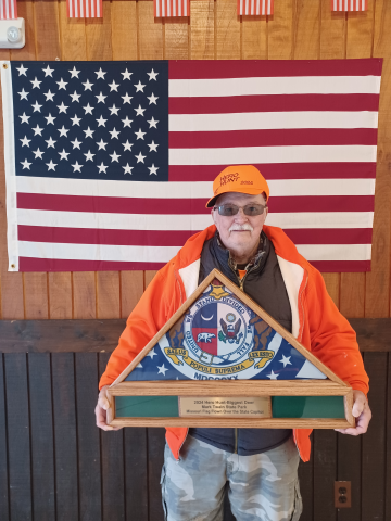 A man standing in front of an American flag holds a plaque labeled 2024 Hero Hunt Biggest Deer