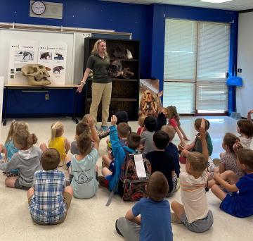 A woman holding a large replica skull speaks to children sitting on a tile floor; many of them have their hands raised