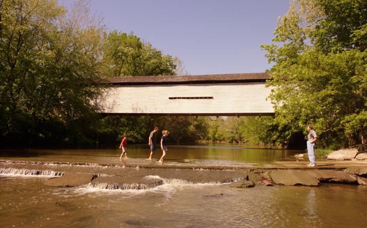 A family walking across a shallow river in front of a covered bridge