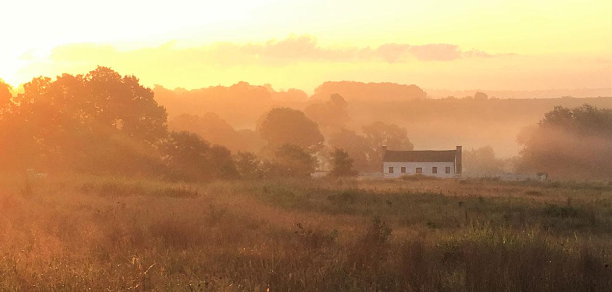 Sunrays gleam across a grassland with a black and white log cabin in the background