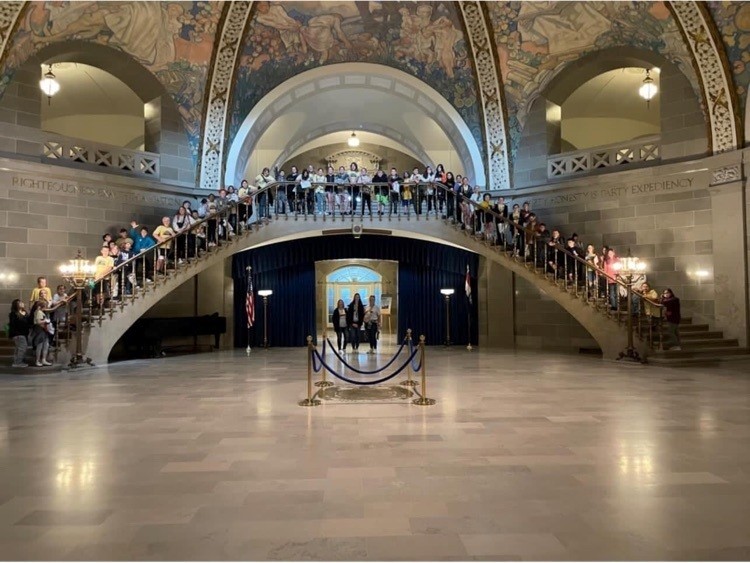 Students stand on the arched stairway of the Capitol rotunda, while  three adults stand on the floor, centered between the two ends of the stairway.