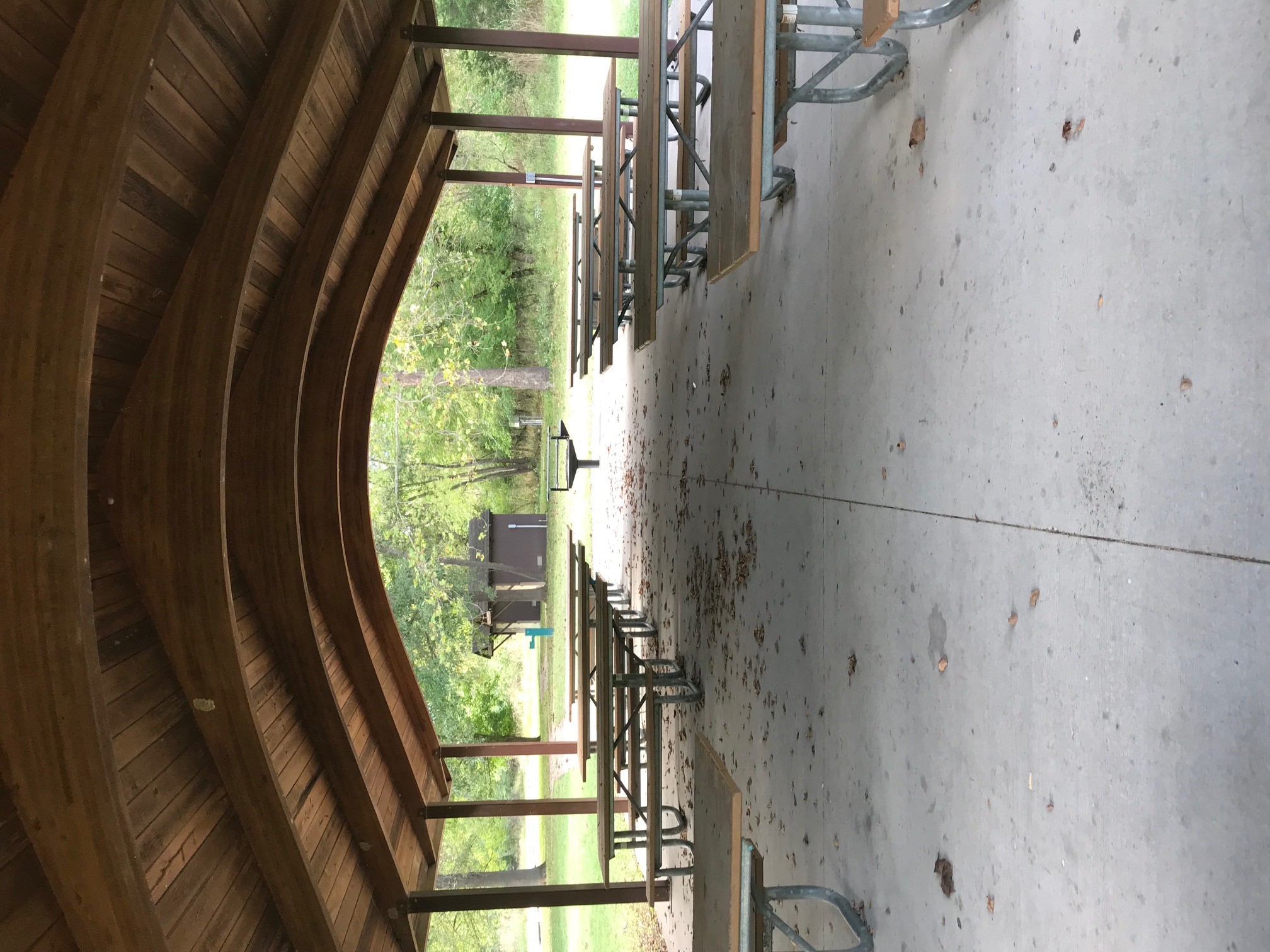 Two rows of picnic tables under the roof of the open shelter, with a grill, water fountain and restroom in the backcground