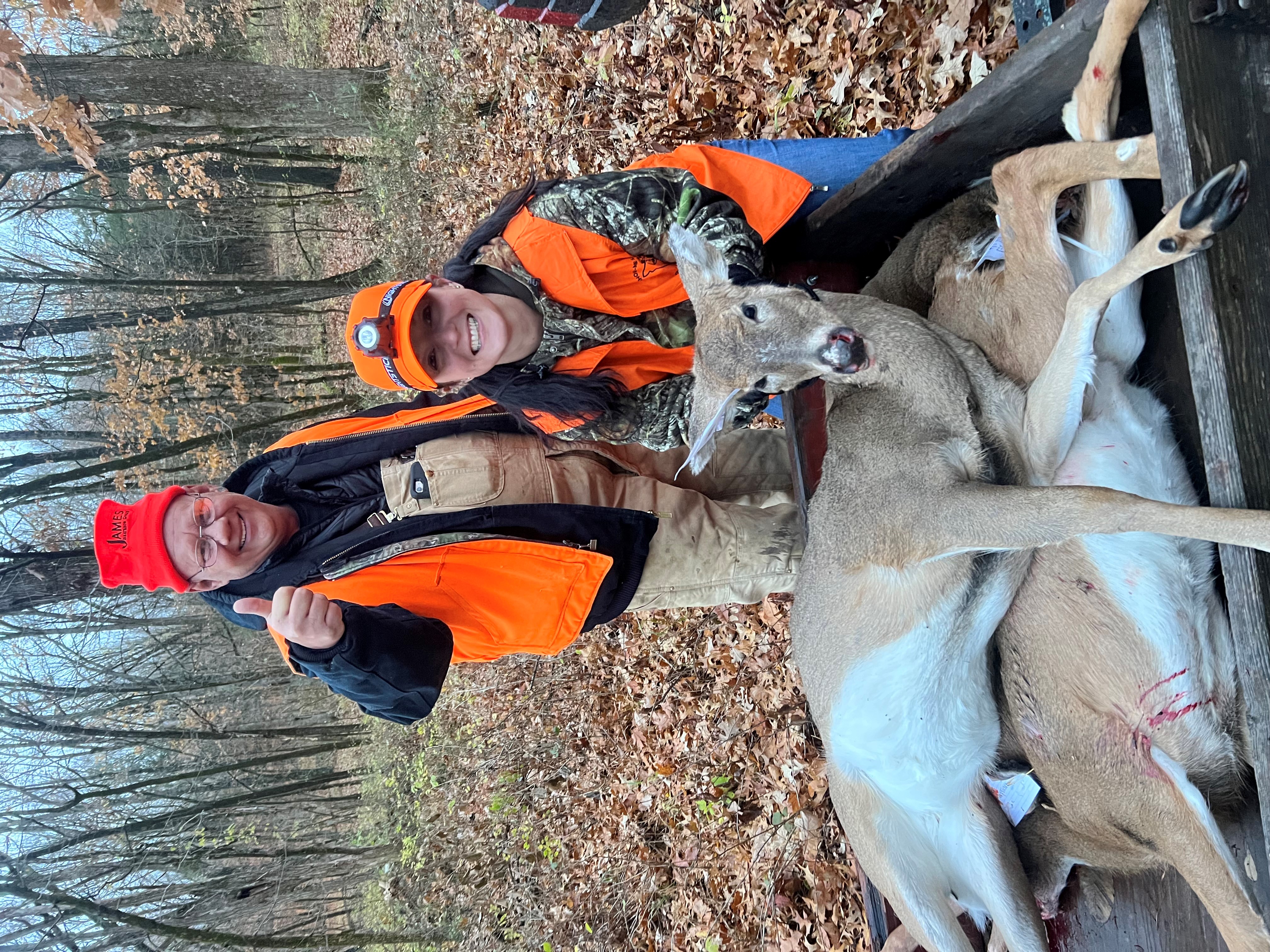A veteran and a guide, both in camouflage and hunter orange pose with a harvested buck in the woods. 
