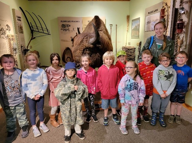 Eleven students and a teacher stand in front of exhibits, including a stuffed bison.