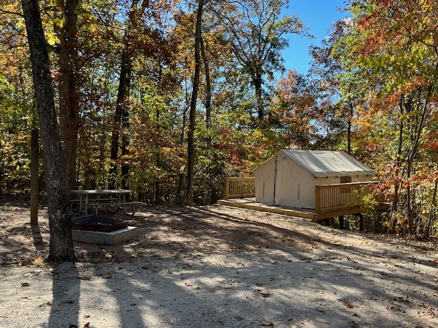 A closed beige-colored canvas tent sits on a wooden deck in a sloping area. The deck is surrounded on three sides by a wooden fence and is supported in the back by posts. A picnic table and fire pit ring in front of the deck, and trees with leaves of red, yellow and green tower over the tent., providing patches of shade.