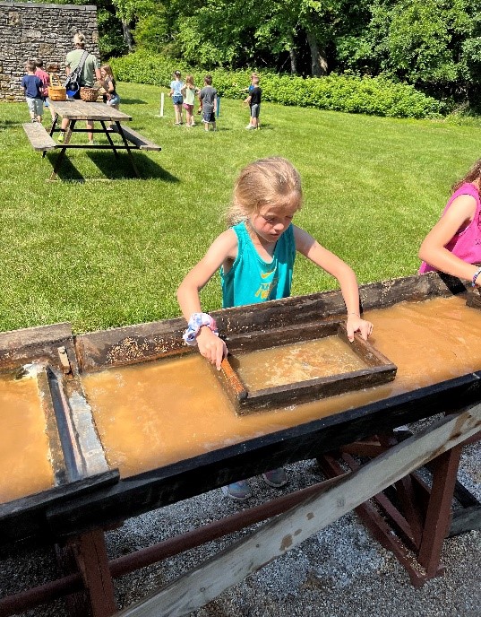 Two girls pan for gems using sieves in a sluice while other students play cornhole and ring toss in the background.