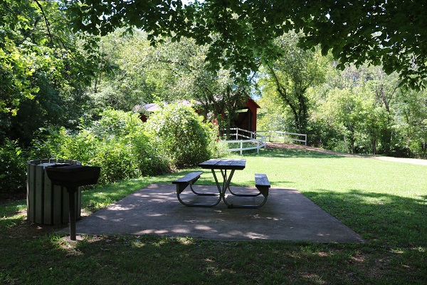 a shaded picnic table and grill with the covered bridge in the background
