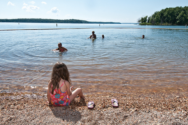 a little girl sitting on the beach watching other kids swim