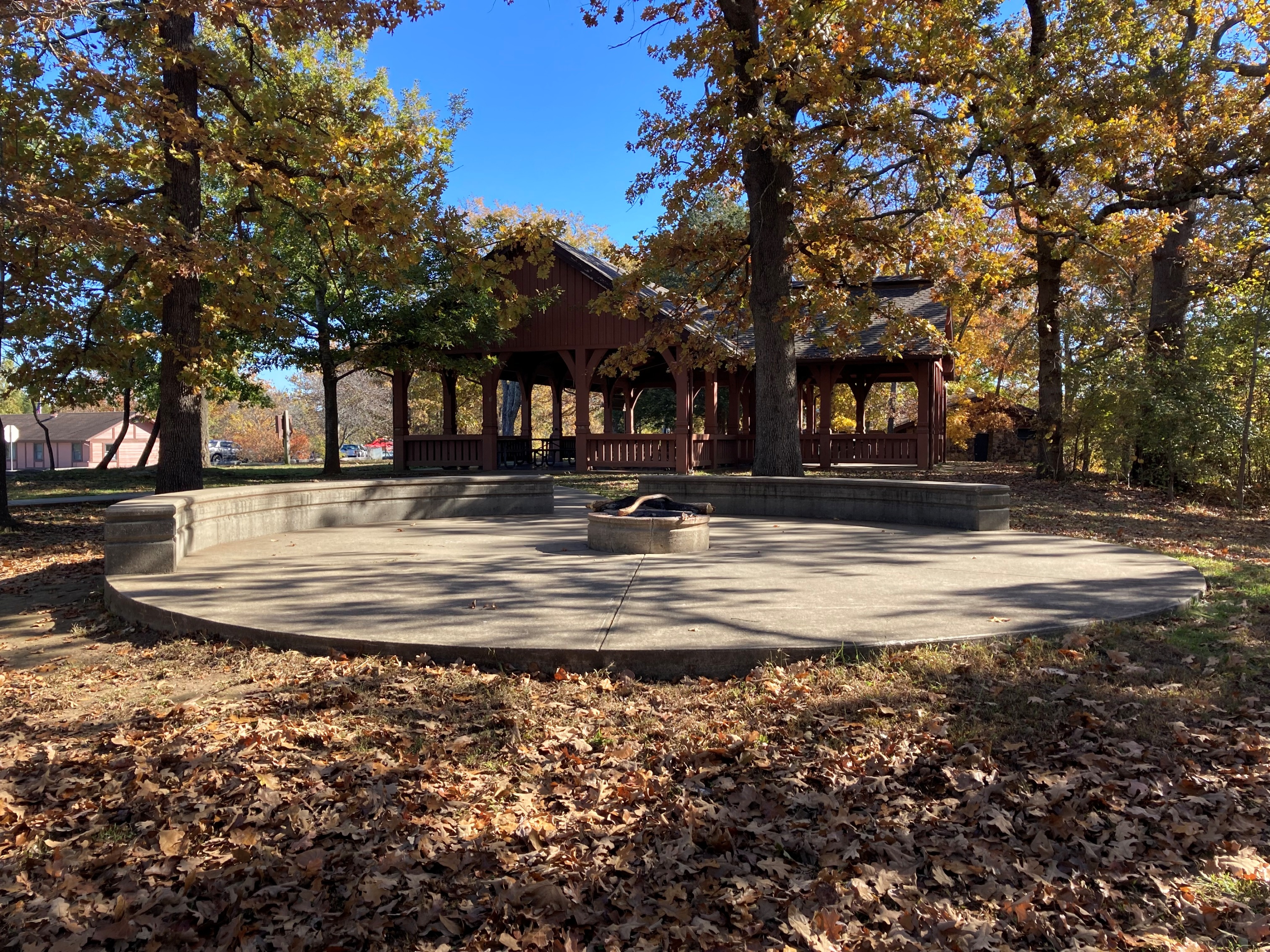 A fire pit sits on a large, circular concrete area near the open picnic shelter.