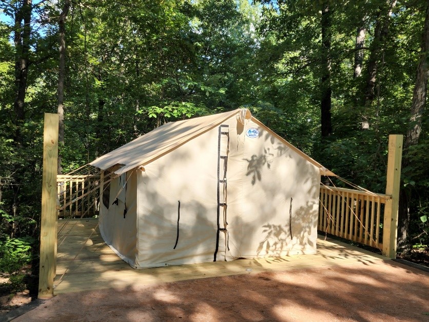 A closed beige-colored tent sits on a wooden platform bordered on three sides by a wooden fence, to which the tent is attached by ropes. Surrounding the fence are trees covered in green foliage, which cast shadows on the tent. 