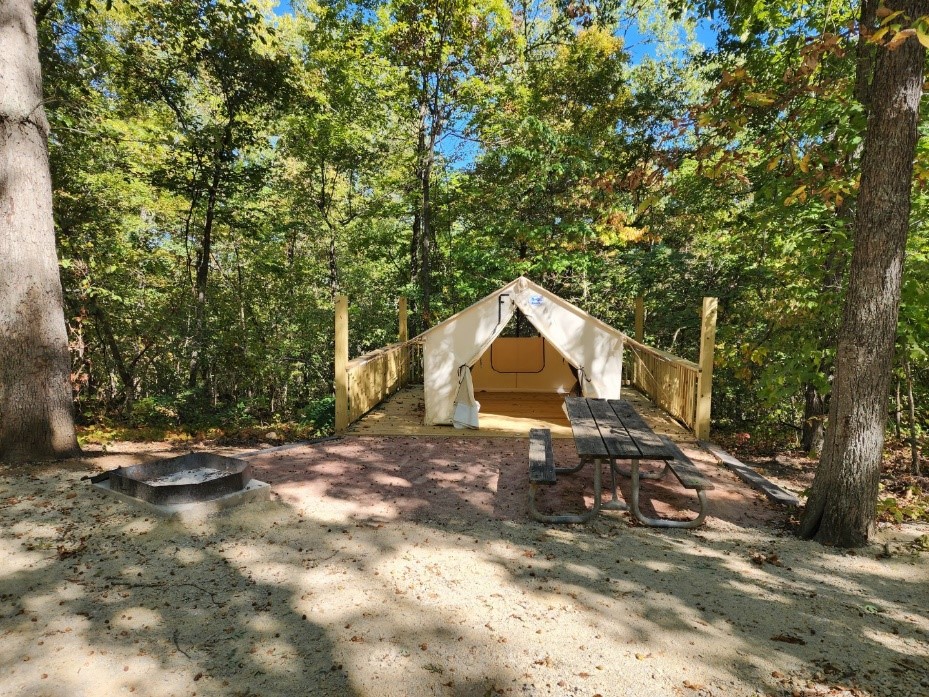 An open beige-colored tent sits on a wooden platform in a shady area surrounded by trees. On the ground in front of the tent are a wooden picnic table and shallow fire ring.