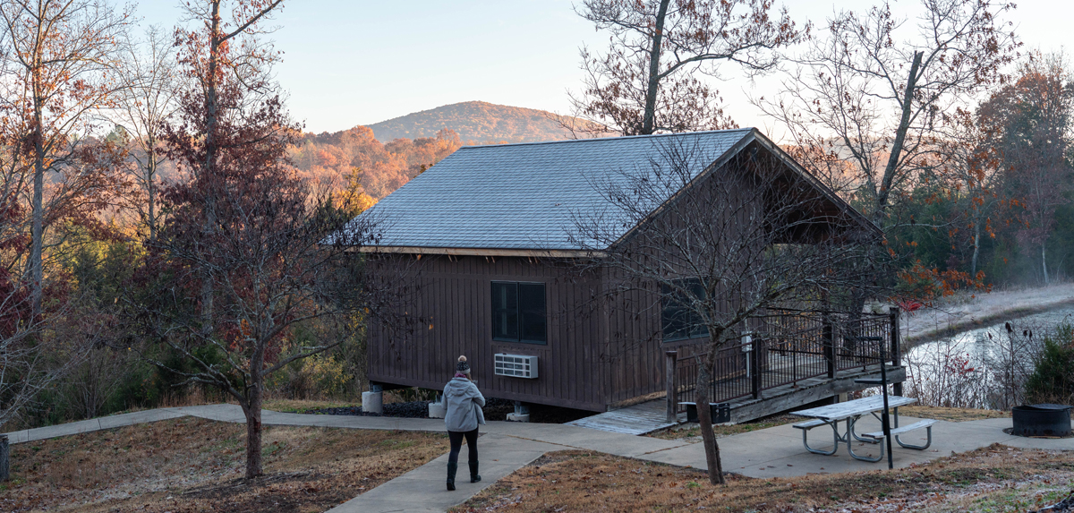 Cabin with surrounding trees and person walking on sidewalk in front of cabin.